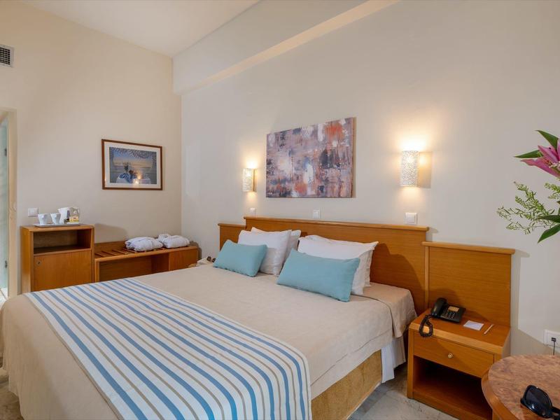 Lichte hotelkamer met groot tweepersoonsbed, blauw gestreept beddengoed en bloemendecoratie.