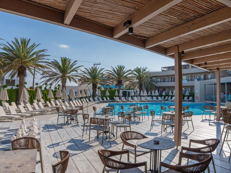 Groot hotelzwembad met ligstoelen en tafels onder houten overkapping bij zonnig weer.