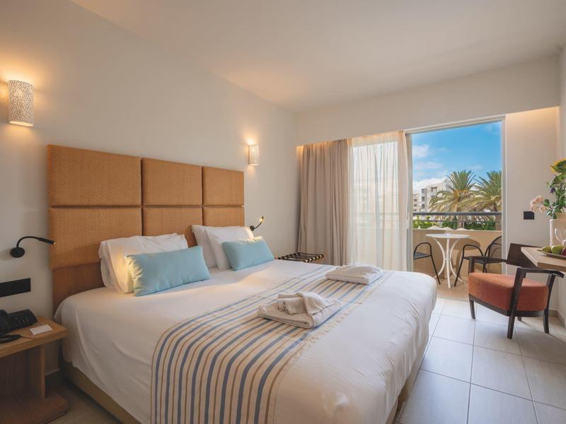 Lichte hotelkamer met groot bed, balkon en uitzicht op palmbomen.