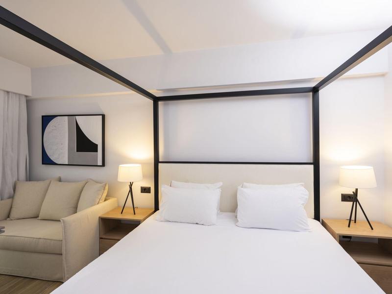 Camera d'hotel moderna con letto baldacchino, divano e due lampade da comodino in tonalità neutre.