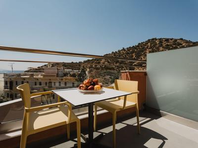 Balkon mit zwei Stühlen und einem Tisch mit Obstschale, mit Blick auf Gebäude und Hügel.