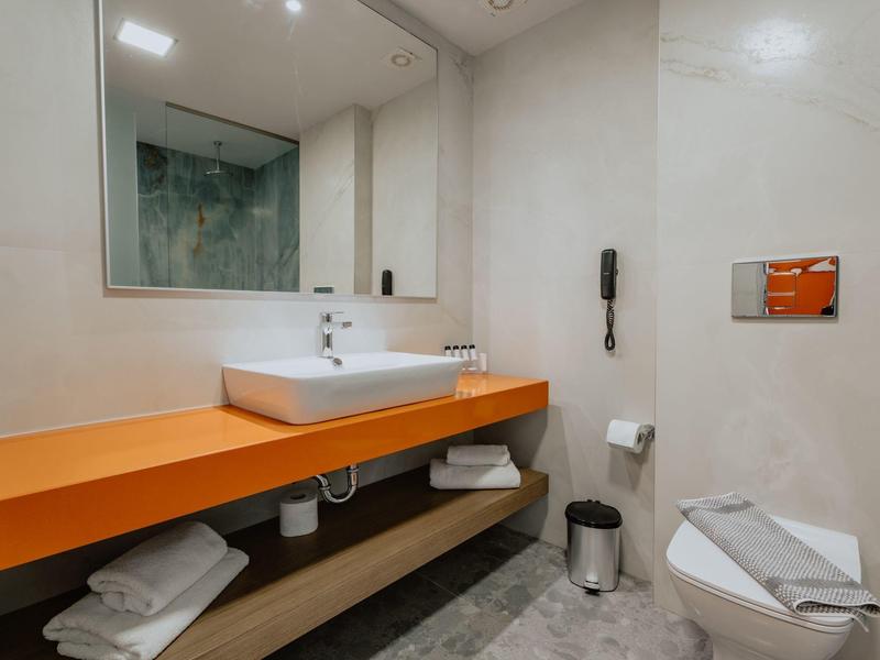 Moderní koupelna s oranžovým umyvadlem a zrcadlem, vybavená ručníky a toaletou.
