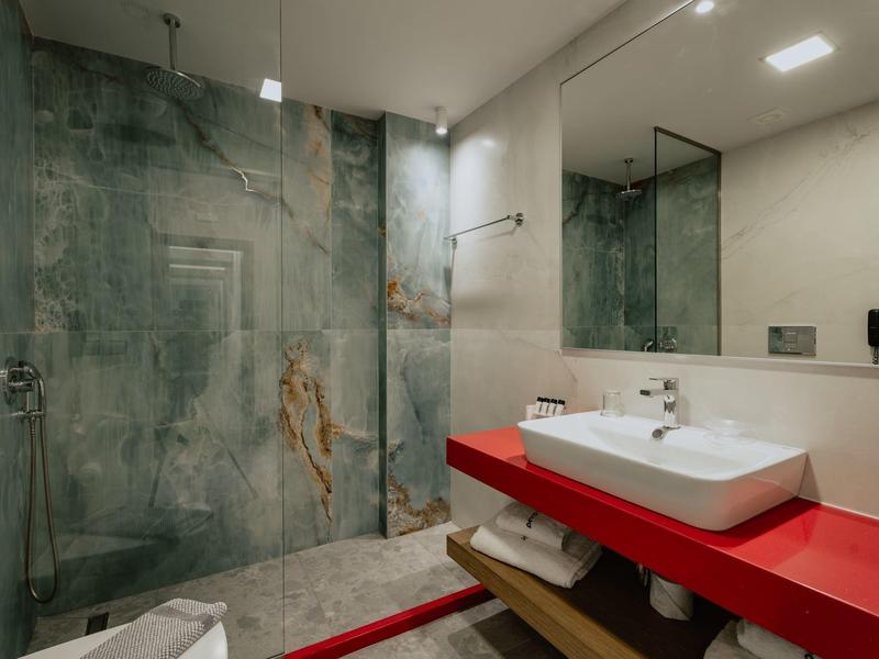 Moderní koupelna s červenou umyvadlovou skříňkou, velkým zrcadlem a skleněným sprchovým koutem s mramorovou stěnou.