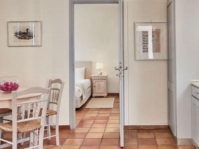 Cozy dining area with wooden chairs and table, bright kitchen with white cabinets, bedroom in background.