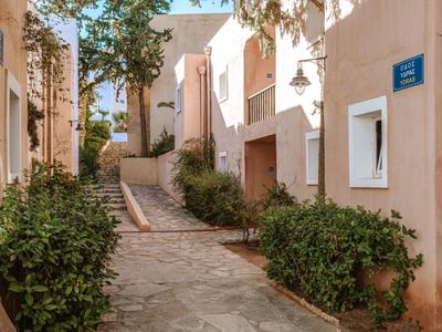 Charming stone-paved alley with trees and plants between light-colored buildings.