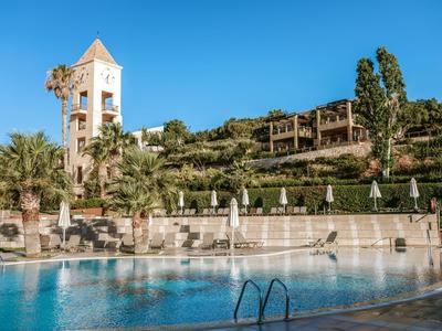 Hôtel avec jardin, piscine et tour sous un ciel bleu clair.