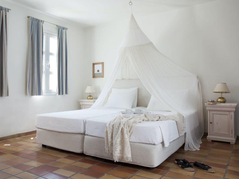 Bright bedroom with white bed, mosquito net, and nightstands beside two windows.