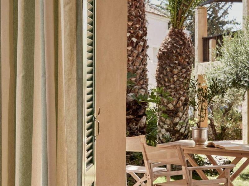 View of a garden with palm trees and chairs through beige curtains in a cozy room.