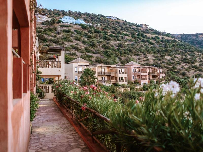 Path beside red buildings with green vegetation and hills in the background.