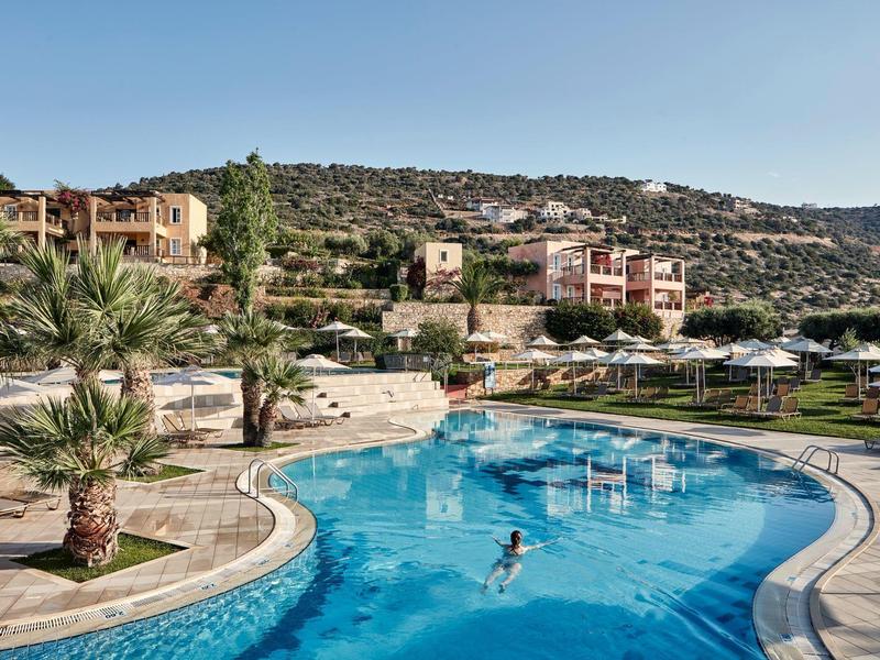 Curved outdoor pool with swimmers, surrounded by palm trees and hillside buildings under clear sky.
