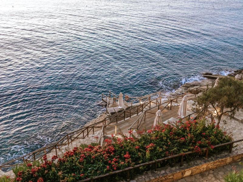 Coastal stairs descend to clear blue sea with flowers and trees nearby.