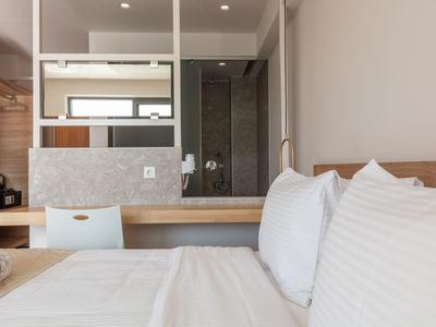 Prosty pokój hotelowy z łóżkiem, drewnianymi detalami i otwartą łazienką z szklaną ścianą.