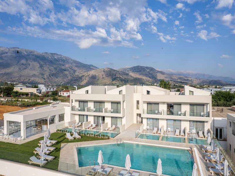 Nowoczesny kompleks hotelowy z basenem i leżakami na tle gór i niebieskiego nieba.