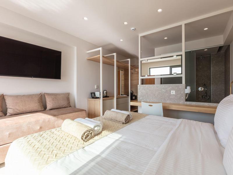 Jasny pokój hotelowy z łóżkiem podwójnym, sofą, telewizorem i nowoczesną szklaną wanną.