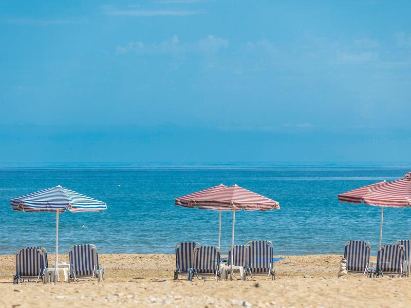 Piaszczysta plaża z trzema leżakami i parasolami na tle spokojnego, niebieskiego morza.