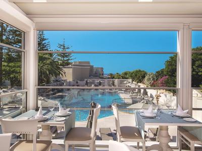 Blick von einem Restaurant mit gedeckten Tischen auf einen Pool und Bäume bei klarem Himmel.