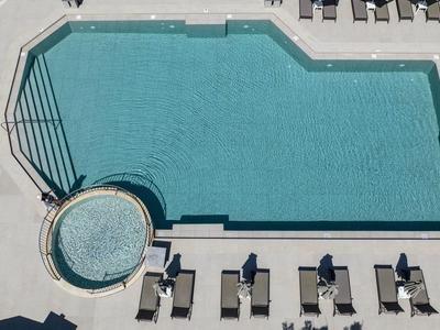 Blick von oben auf einen rechteckigen Pool mit angrenzendem rundem Whirlpool und Liegestühlen auf einer hellen Terrasse.