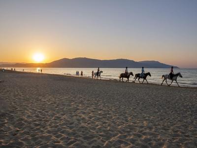 Západ slunce na pláži s lidmi a třemi koňmi kráčejícími podél pobřeží