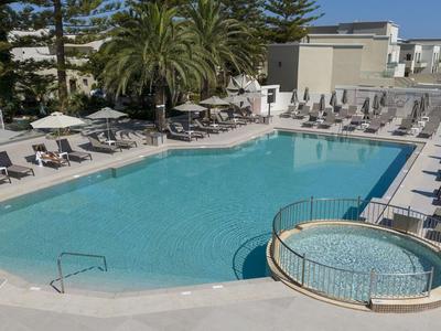 Una piscina all'aperto con lettini e palme in un giardino di un hotel sotto un cielo blu.