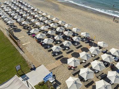 Strand mit vielen Reihen weißer Sonnenschirme und Sonnenliegen, daneben grüner Rasen und Meer.