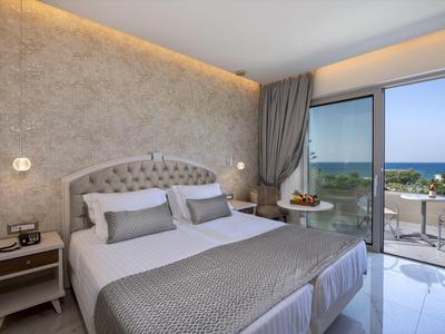 Moderní hotelový pokoj s manželskou postelí, čalouněným čelem a balkonem s výhledem na moře.