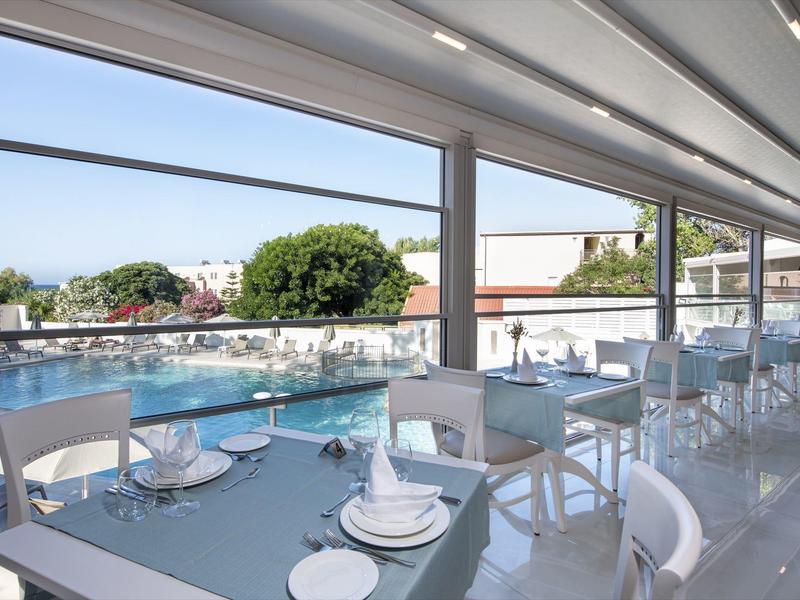 Venkovní jídelní prostor se skleněnými stoly a bílými židlemi s výhledem na bazén a zeleň.