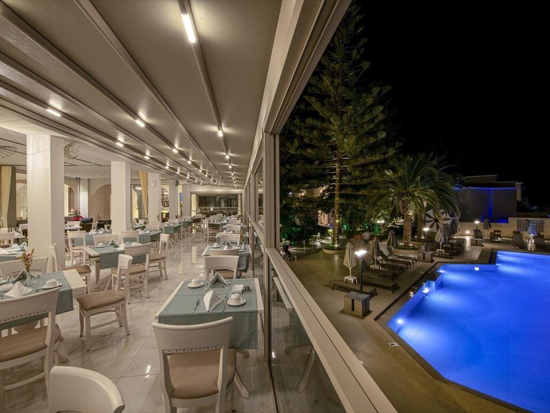 Noční pohled na moderní venkovní jídelní prostor vedle modře osvětleného bazénu a stromů.