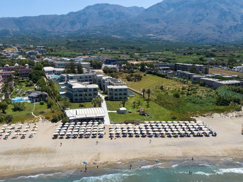 Široká písčitá pláž s řadami lehátek a slunečníků, za nimi zelené trávníky a budovy pod horami.