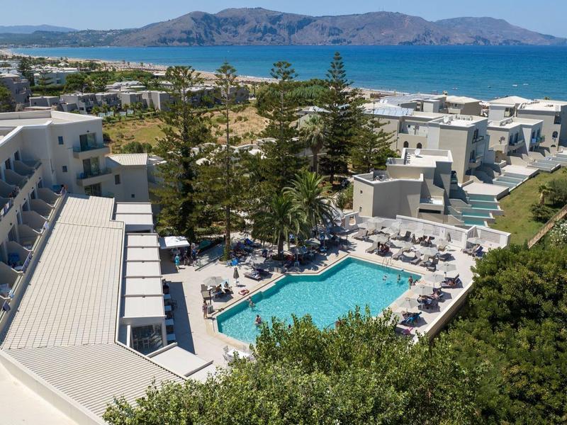 Hotel s bazénem, lehátky, stromy a moře v pozadí pod slunečnou oblohou.