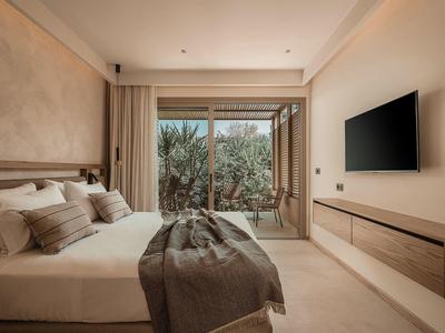 Modernes Schlafzimmer mit großem Bett, Fernseher an der Wand und Blick auf einen Balkon mit Pflanzen.