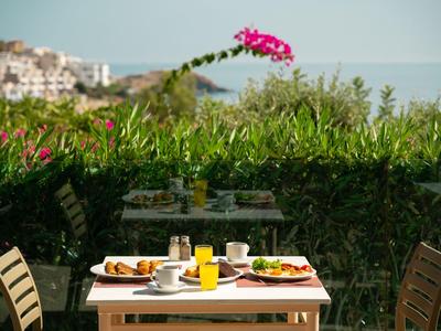 Stół nakryty do śniadania z napojem na tarasie z widokiem na zielone rośliny i białe domy.