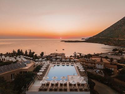 Blick auf einen Pool mit Liegestühlen, dahinter Meer und Berg bei Sonnenuntergang.