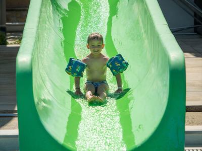 Małe dziecko z rękawkami do pływania zjeżdża z zielonej zjeżdżalni wodnej do wody.