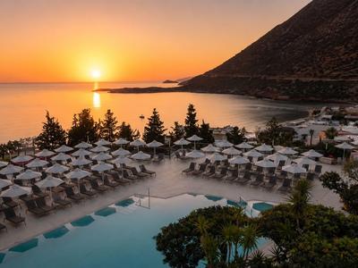 Sonnenuntergang über einem Hotelpool mit weißen Sonnenschirmen am Meer und Bergen im Hintergrund.