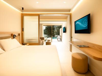 Helles Hotelzimmer mit großem Bett, Wandfernseher und Blick auf Terrasse mit Garten.