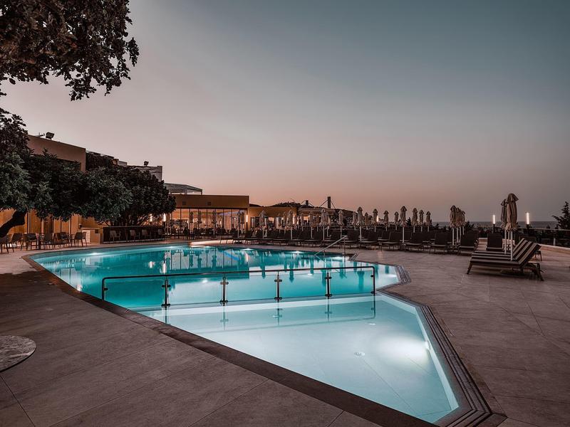 Beleuchteter Außenpool bei Sonnenuntergang mit Liegestühlen und Bäumen im Hintergrund.