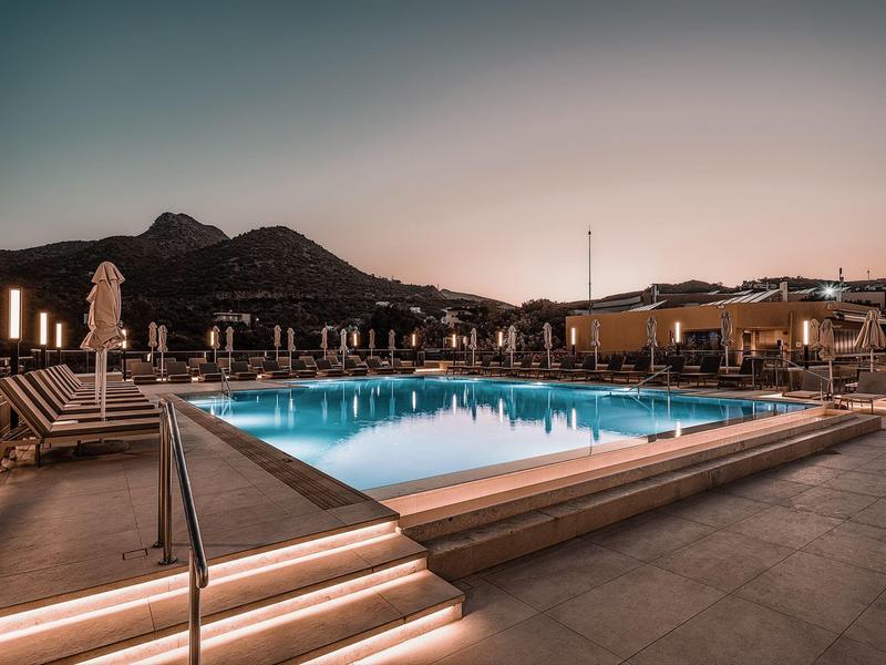 Beleuchteter Außenpool bei Dämmerung mit Liegestühlen und Bergen im Hintergrund.