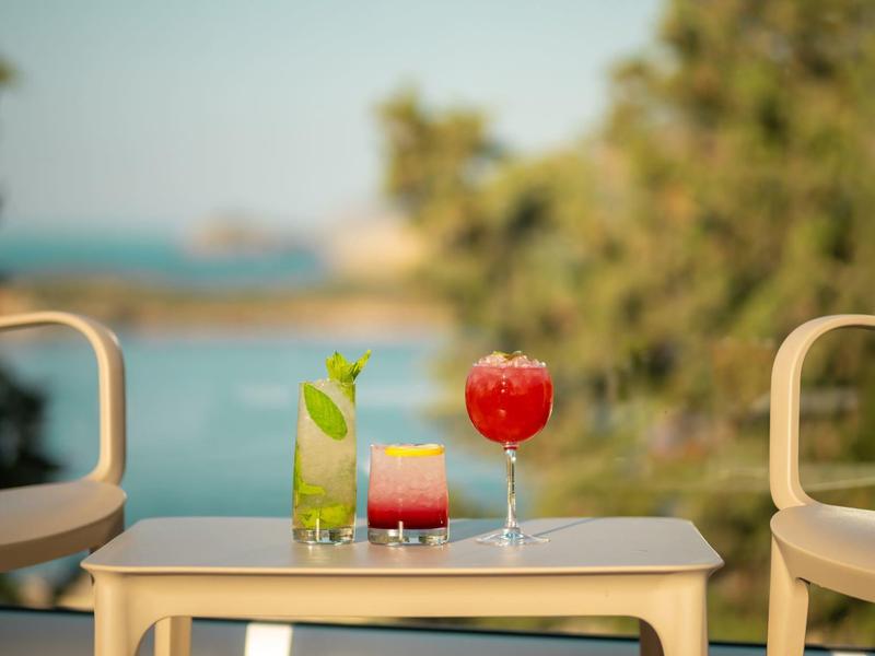 Drei bunte Cocktails auf einem Tisch mit zwei Stühlen vor ruhigem Wasser und grüner Natur.