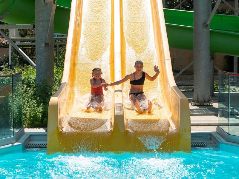 Zwei Kinder rutschen nebeneinander eine gelbe Wasserrutsche ins Schwimmbecken hinunter.