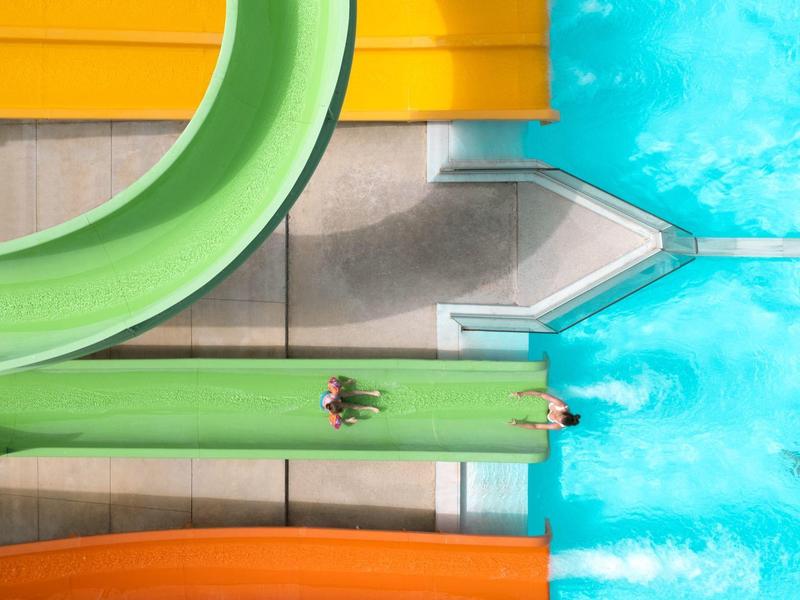 Blick von oben auf Wasserrutschen mit Menschen, die ins blaue Wasser gleiten.