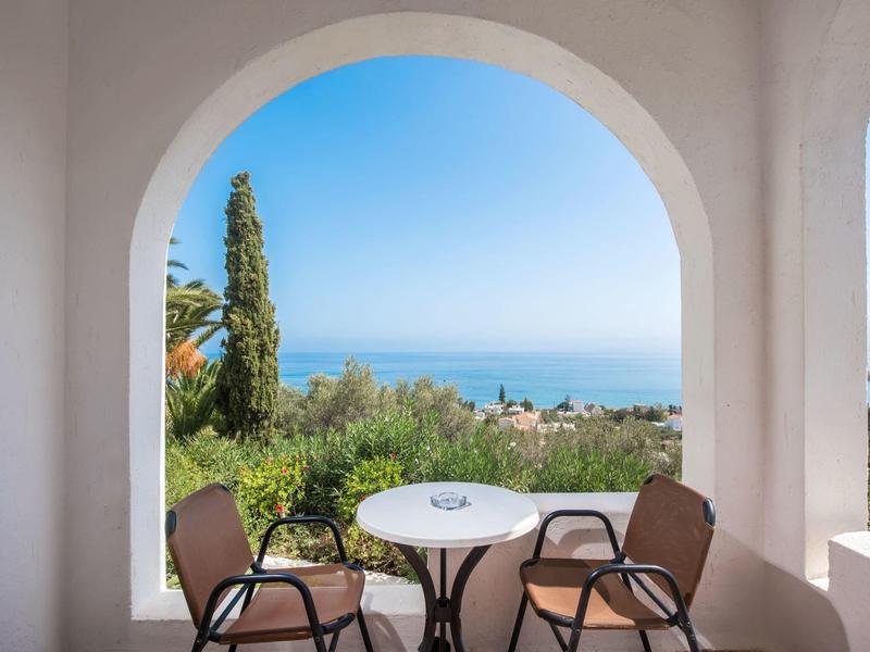 View through a large arch to the sea, garden, and terrace with table and chairs.