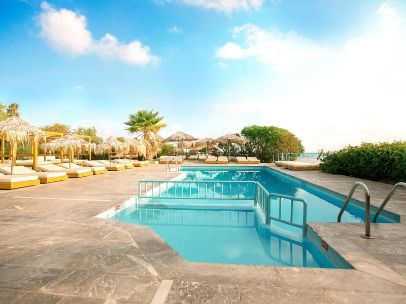 Swimming pool with lounge chairs and umbrellas in a sunny hotel setting overlooking the sea.