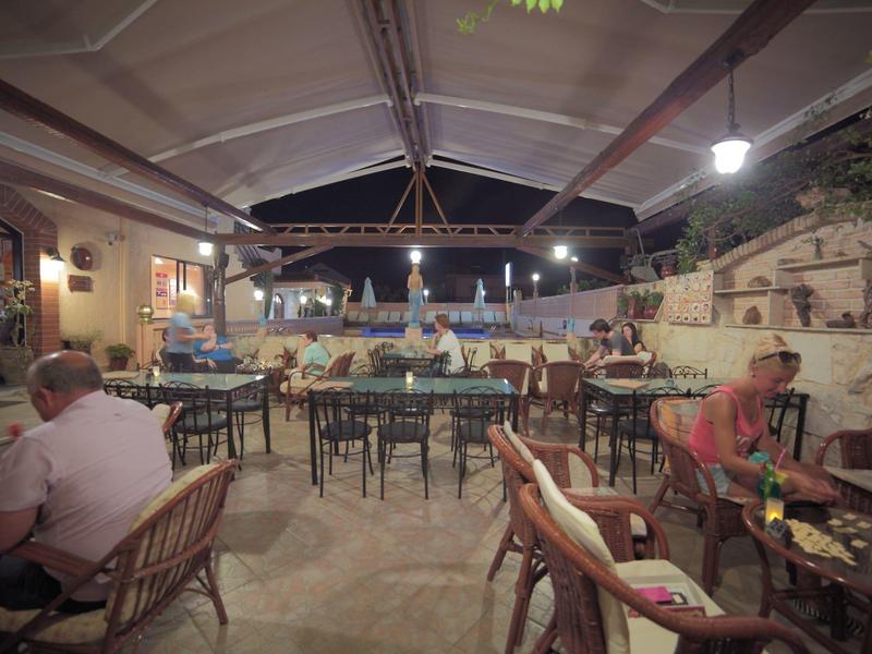 Offene Terrasse mit Holztischen und Stühlen, beleuchtet bei Abenddämmerung, zwei Personen sitzen.