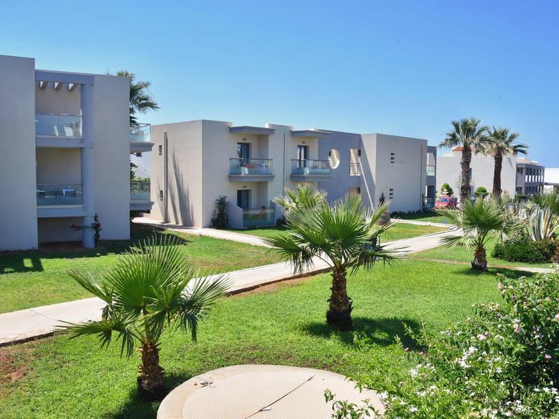 Moderní hotelový komplex s nízkými budovami, travnatými plochami a palmami pod jasnou oblohou.