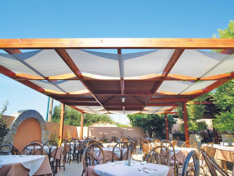 Overdekt buitengedeelte met tafels en stoelen, omgeven door bomen en een heldere lucht.