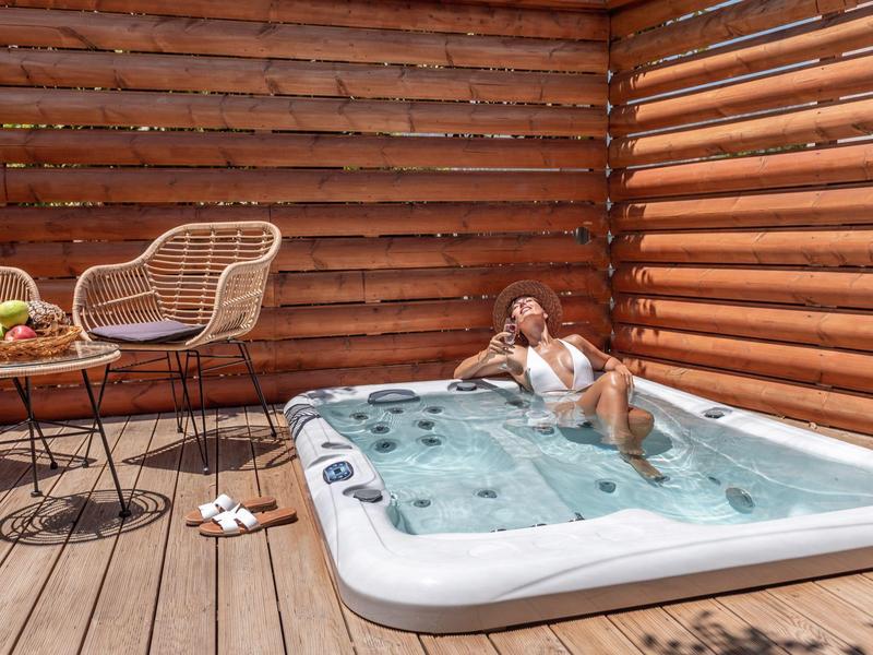 Vrouw ontspant in een jacuzzi op een houten terras naast een tafel met stoelen en een fruitschaal.