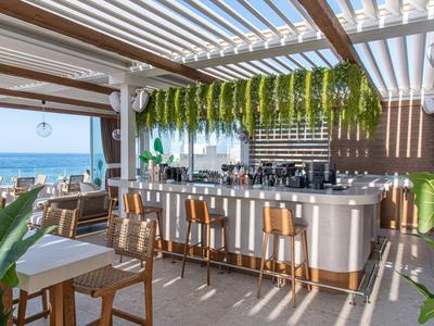 Bar sulla spiaggia aperta con sgabelli alti, bancone e vista mare sotto una pergola.
