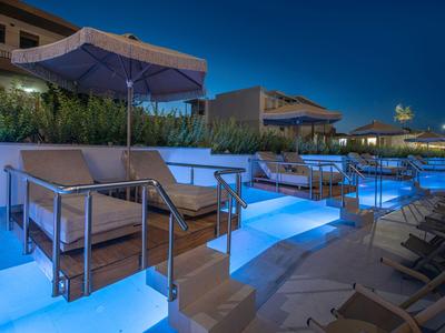 Area piscina illuminata con lettini e ombrelloni davanti a edifici moderni di notte.