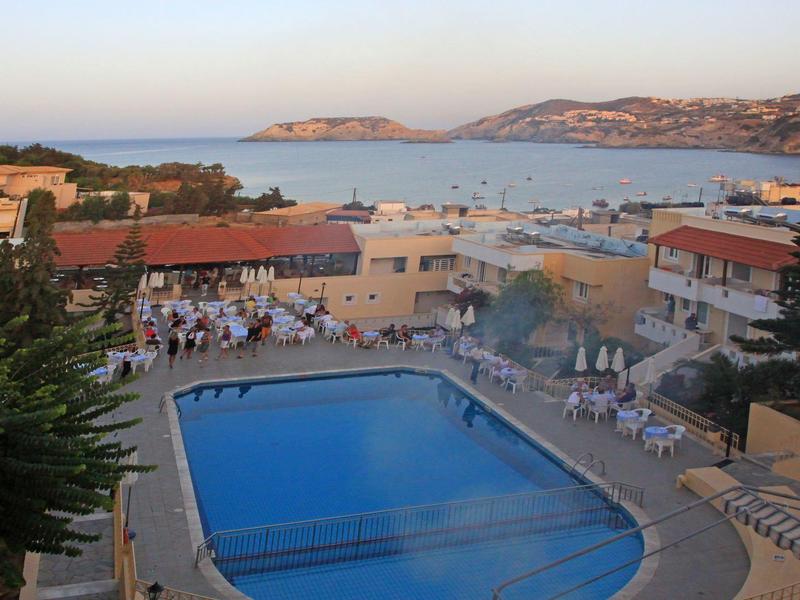 Une piscine d'hôtel avec chaises longues entourée de bâtiments avec vue sur la mer et des collines en arrière-plan.
