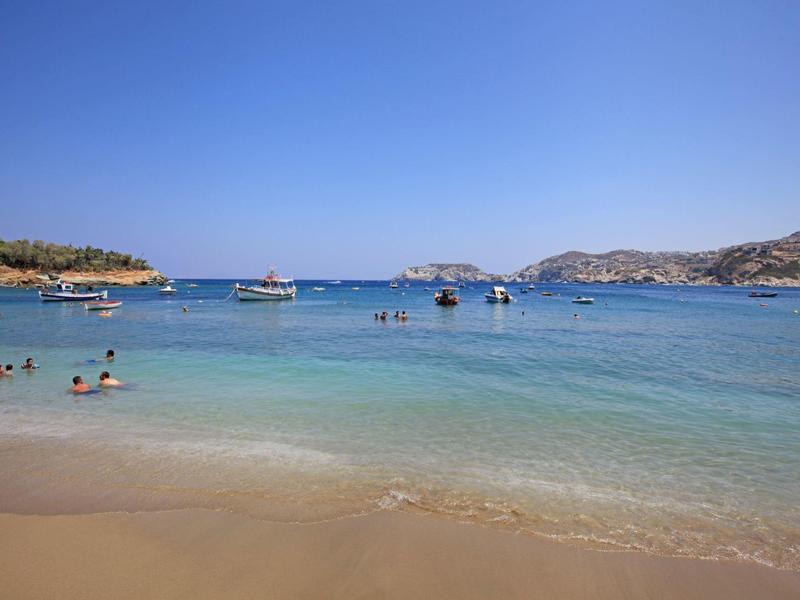 Mer claire avec plage de sable, nageurs et bateaux sous un ciel bleu.